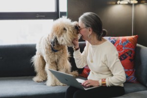 Woman (Honey Good) and her dog on sofa smiling while being licked exuding the luxury of a simple life.