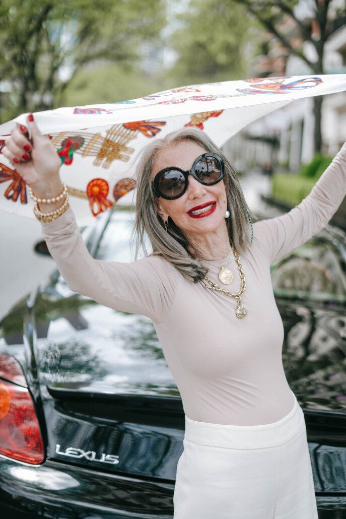 Image of Honey Good out on the street, playfully holding a scarf above her head with sunglasses on a smile as she finds joy in solitary pursuits