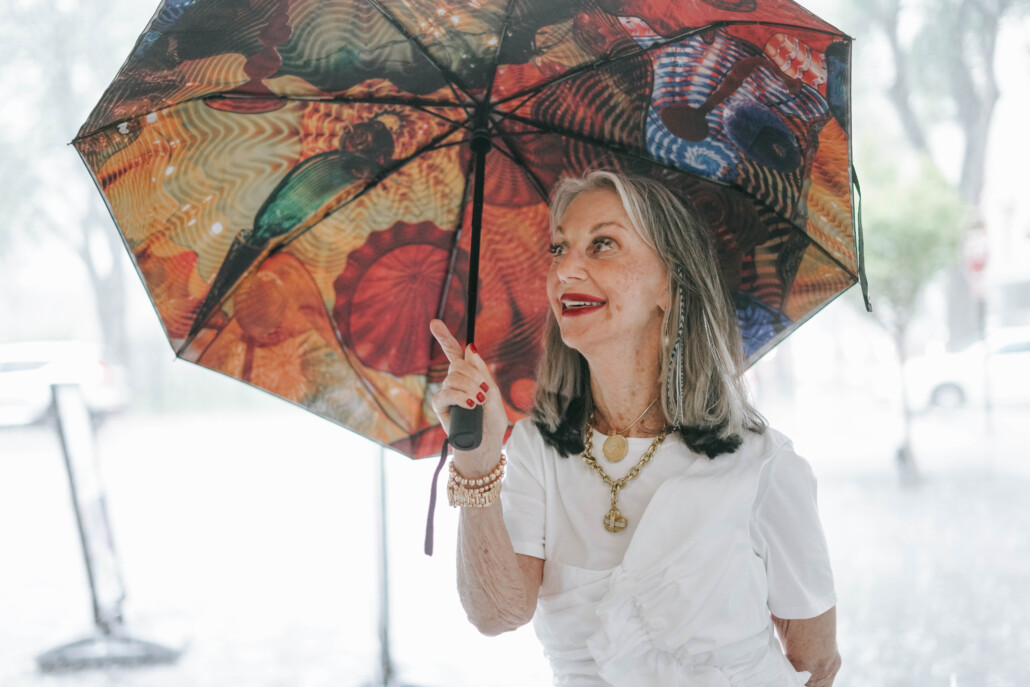 Image of Honey Good holding a colorful umbrella, wearing a white blouse and her signature red lipstick and a smile talking about fashion faux pas for women over 50